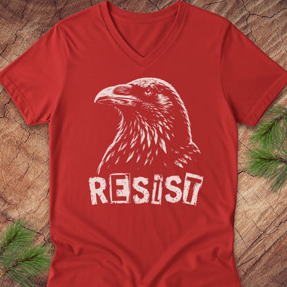 A red V-neck t-shirt featuring a white graphic of a raven and the word 'RESIST' in a distressed stencil font, laid flat on a rustic wood background with pine needles.