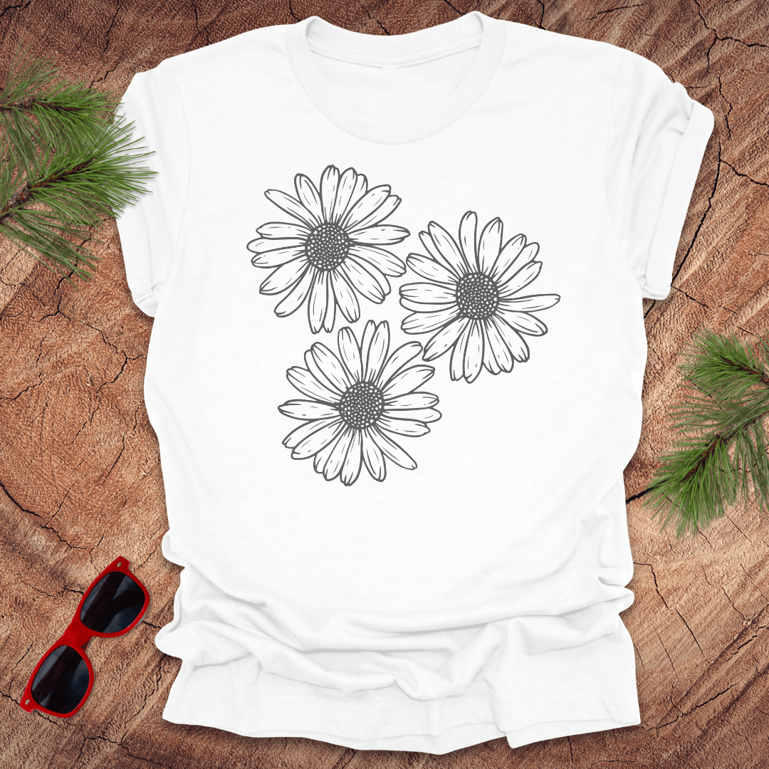 A white t-shirt with 3 daisies prominently displayed. on a wooden surface with sunglasses and greenery.