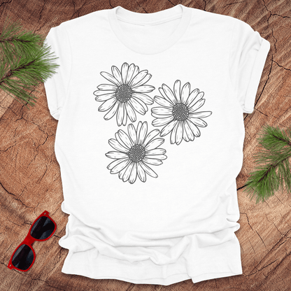 A white t-shirt with 3 daisies prominently displayed. on a wooden surface with sunglasses and greenery.