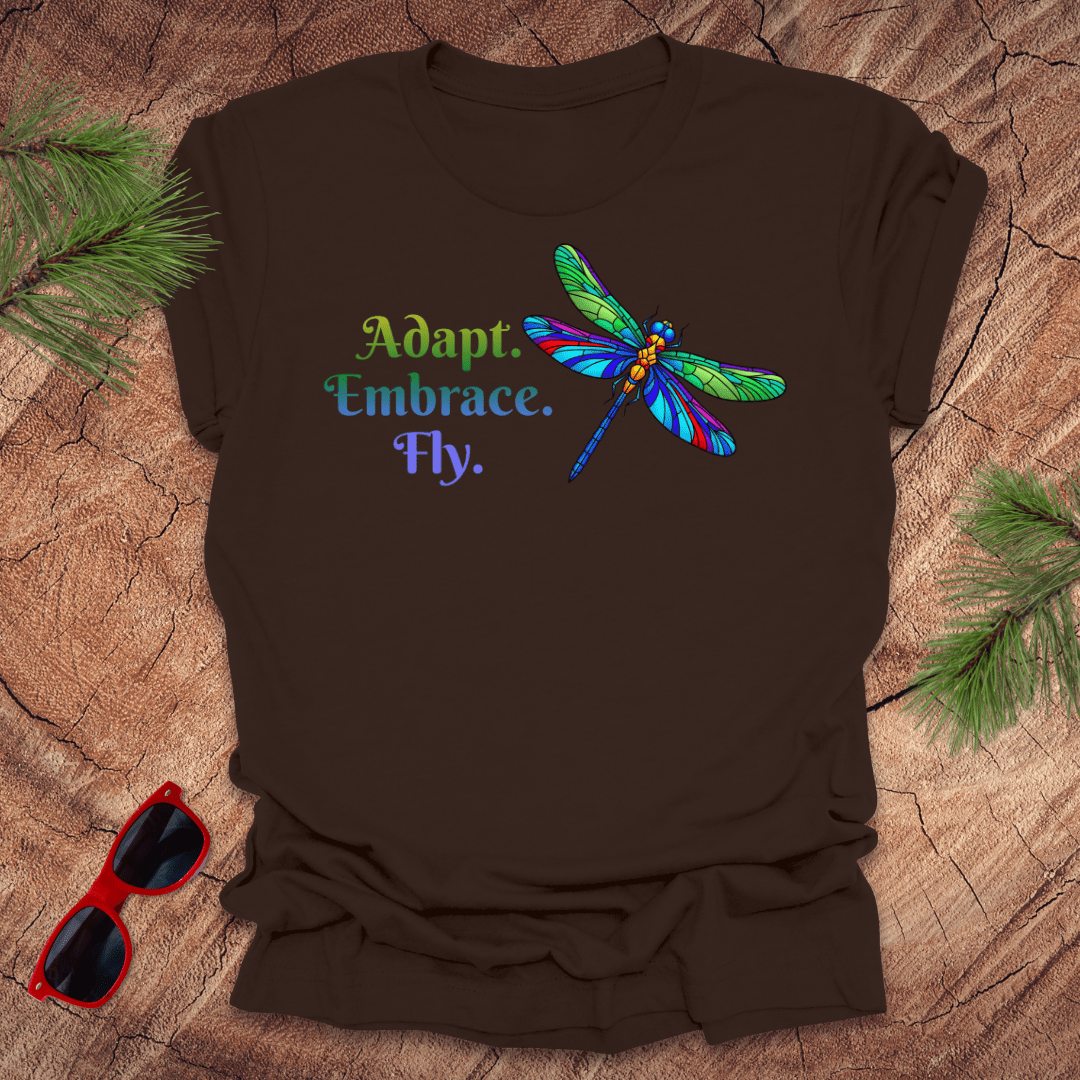 Brown t-shirt with colorful dragonfly graphic and text on a wooden surface with sunglasses and pine branches.