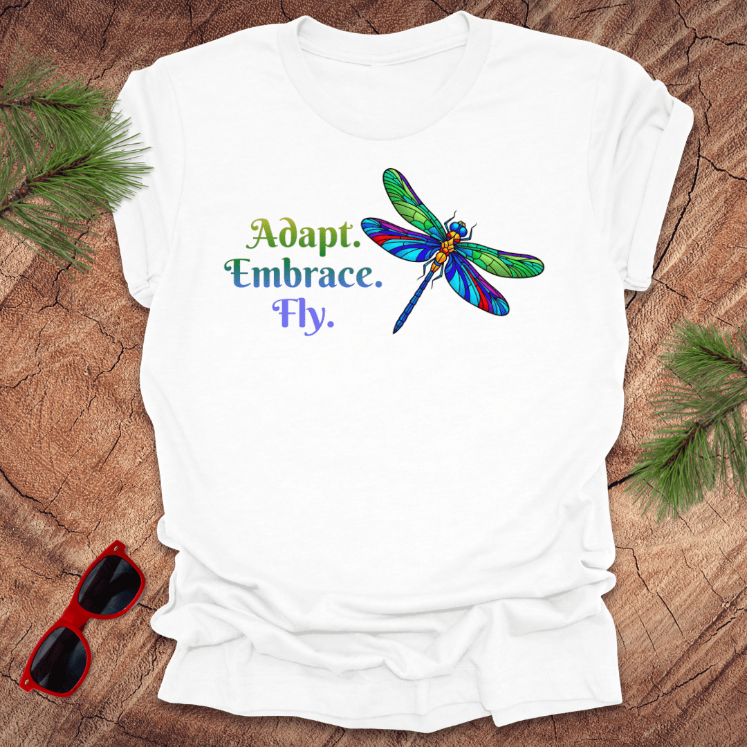 White t-shirt with colorful dragonfly graphic and text 'Adapt. Embrace. Fly on a wood background