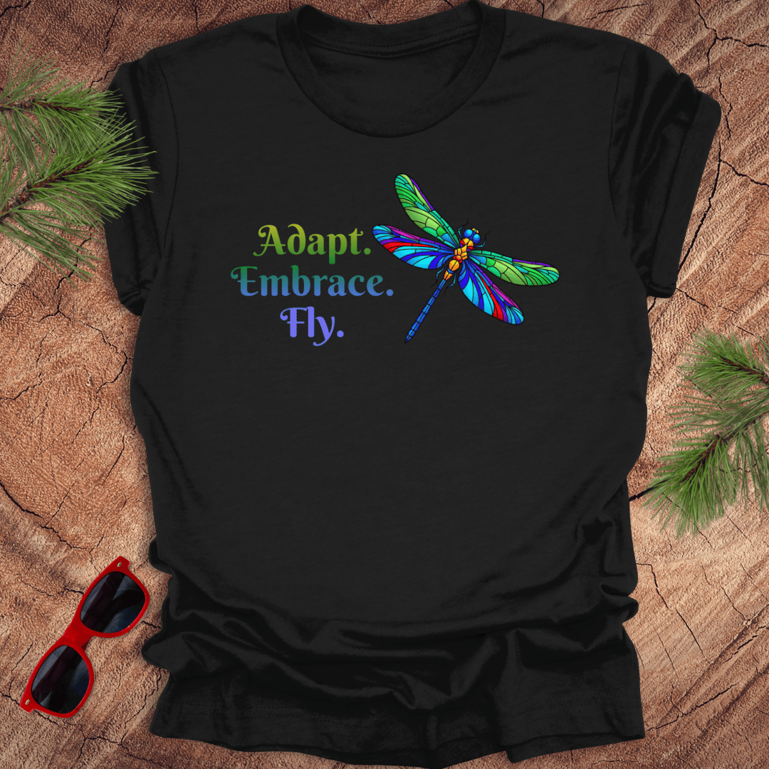 Black t-shirt with colorful dragonfly design and text on a wooden surface with sunglasses and pine branches.