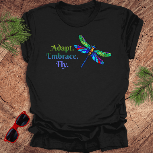 Black t-shirt with colorful dragonfly design and text on a wooden surface with sunglasses and pine branches.