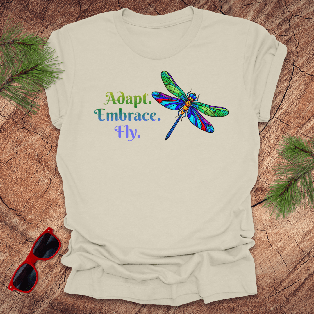 Beige t-shirt with colorful dragonfly graphic and text on a wooden surface with sunglasses and greenery.
