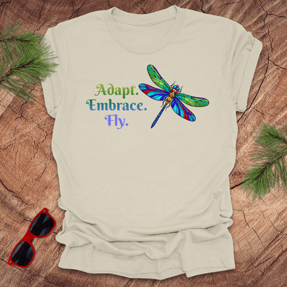 Beige t-shirt with colorful dragonfly graphic and text on a wooden surface with sunglasses and greenery.