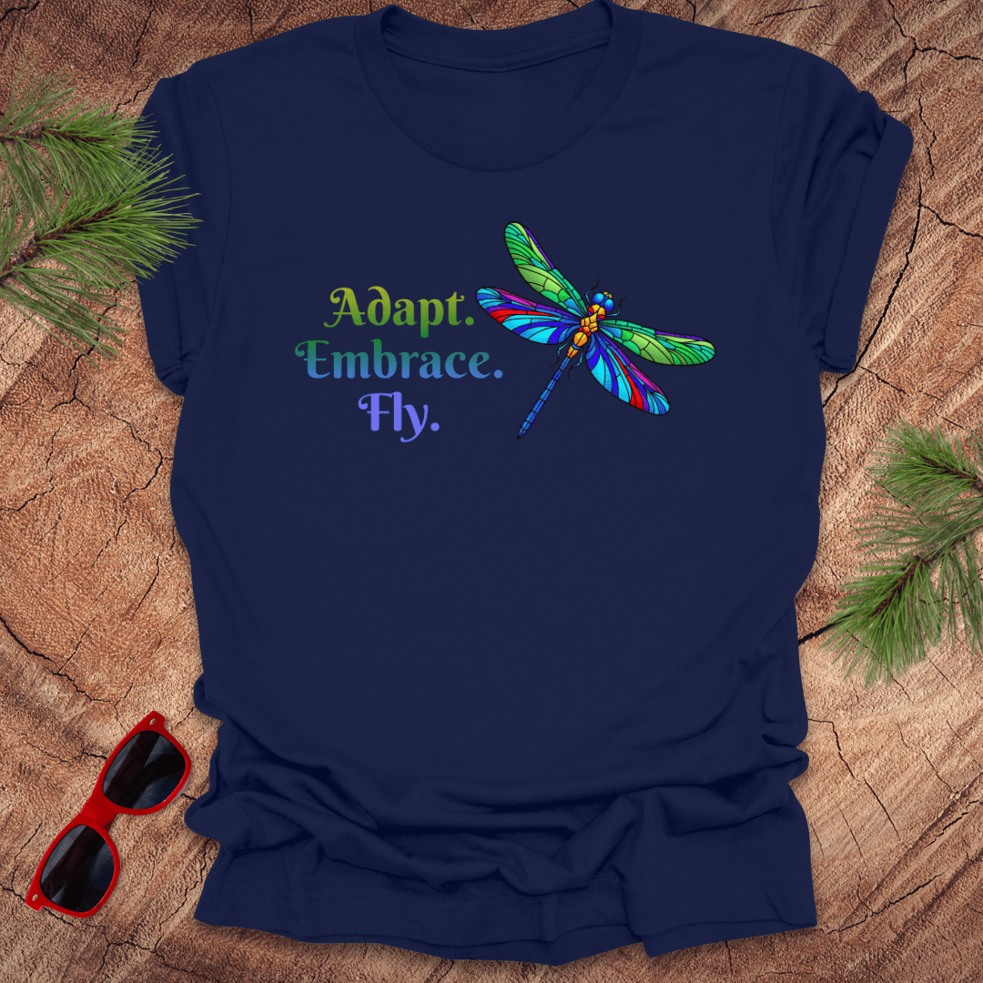 Navy blue t-shirt with colorful dragonfly graphic and text on a wooden surface with sunglasses and greenery.