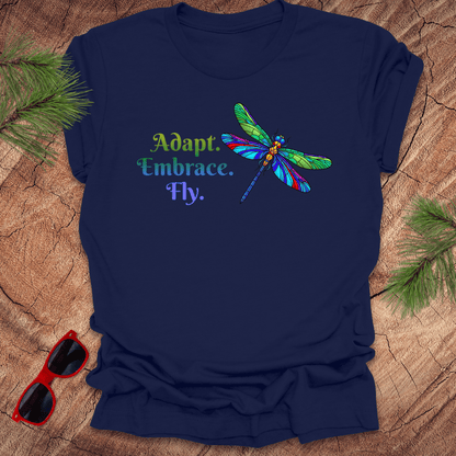Navy blue t-shirt with colorful dragonfly graphic and text on a wooden surface with sunglasses and greenery.