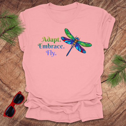 Pink t-shirt with a colorful dragonfly graphic and text on a wooden surface with sunglasses and greenery.