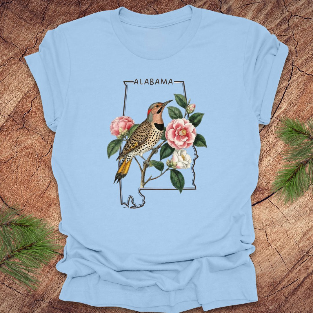 Baby blue t-shirt with Alabama state outline design featuring a northern flicker and camellia flowers on a wooden surface.