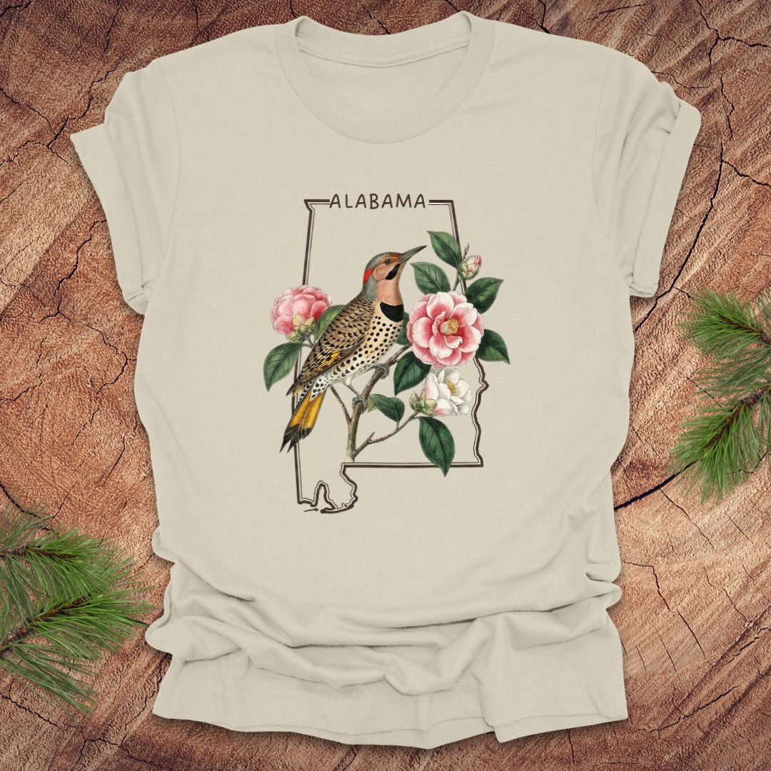 Beige t-shirt with Alabama state outline design featuring a northern flicker and camellia flowers on a wooden surface.