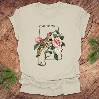 Beige t-shirt with Alabama state outline design featuring a northern flicker and camellia flowers on a wooden surface.