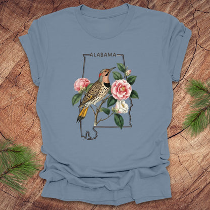 Steel blue t-shirt with Alabama state outline design featuring a northern flicker and camellia flowers on a wooden surface.