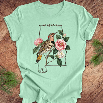 Mint green t-shirt with Alabama state outline design featuring a northern flicker and camellia flowers on a wooden surface.
