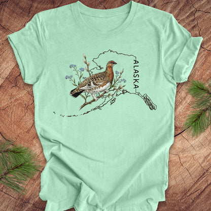 Mint green t-shirt with Alaska state outline featuring a ptarmigan on a wooden surface.