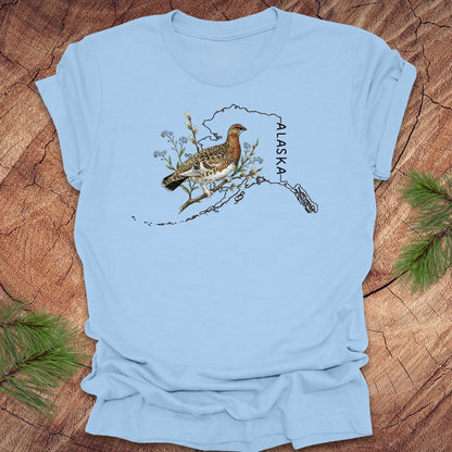 Baby blue t-shirt with Alaska state outline featuring a ptarmigan on a wooden surface.