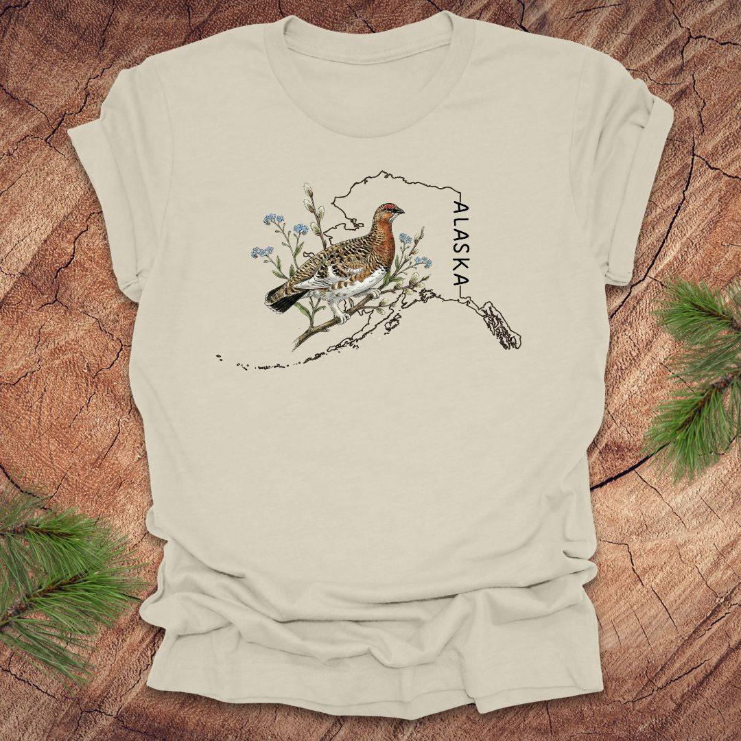 Beige t-shirt with Alaska state outline featuring a ptarmigan on a wooden surface.