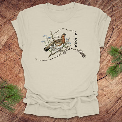 Beige t-shirt with Alaska state outline featuring a ptarmigan on a wooden surface.