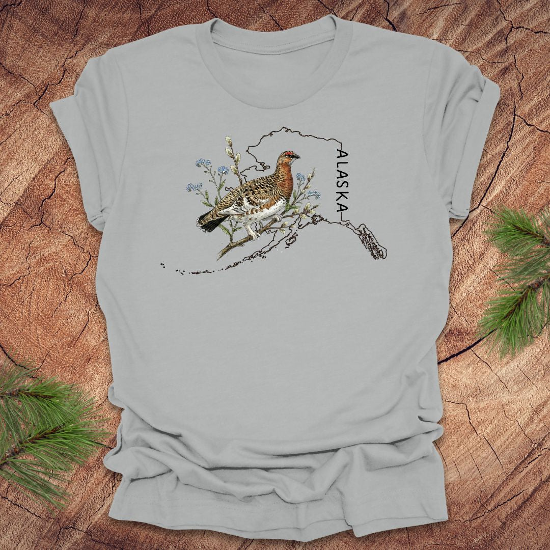 Silver t-shirt with Alaska state outline featuring a ptarmigan on a wooden surface.