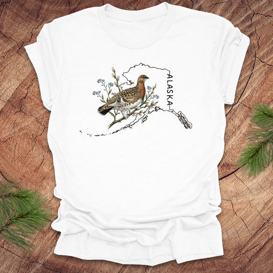 White t-shirt with Alaska state outline featuring a ptarmigan on a wooden surface.