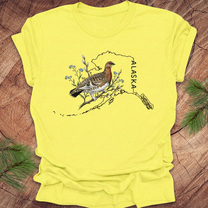 Yellow t-shirt with an Alaska-themed graphic featuring a bird and nature elements on a wooden surface.
