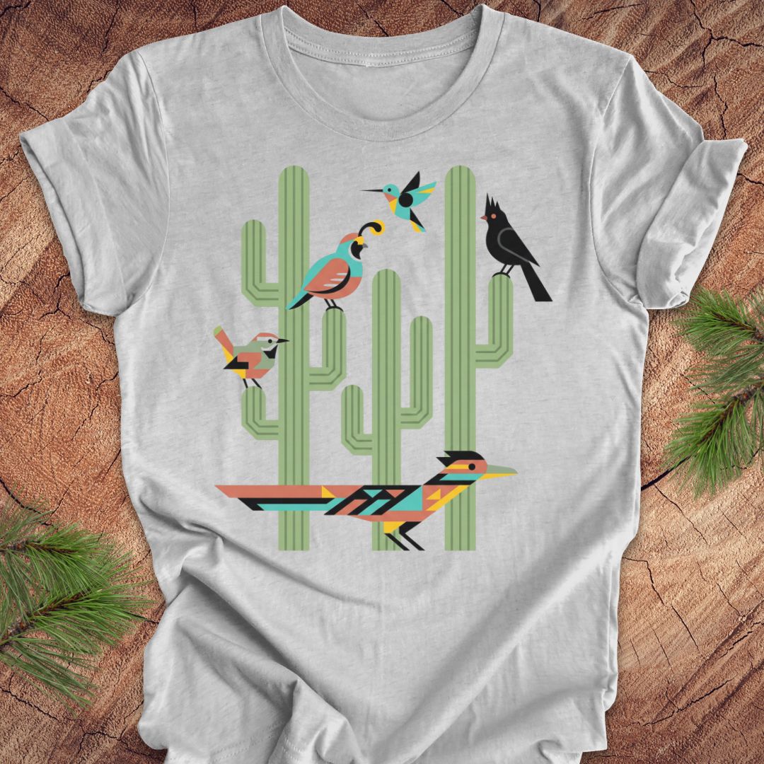 A heather gray t-shirt with a colorful graphic print of stylized birds perched on green cacti, laid flat on a rustic wood background with pine needles.