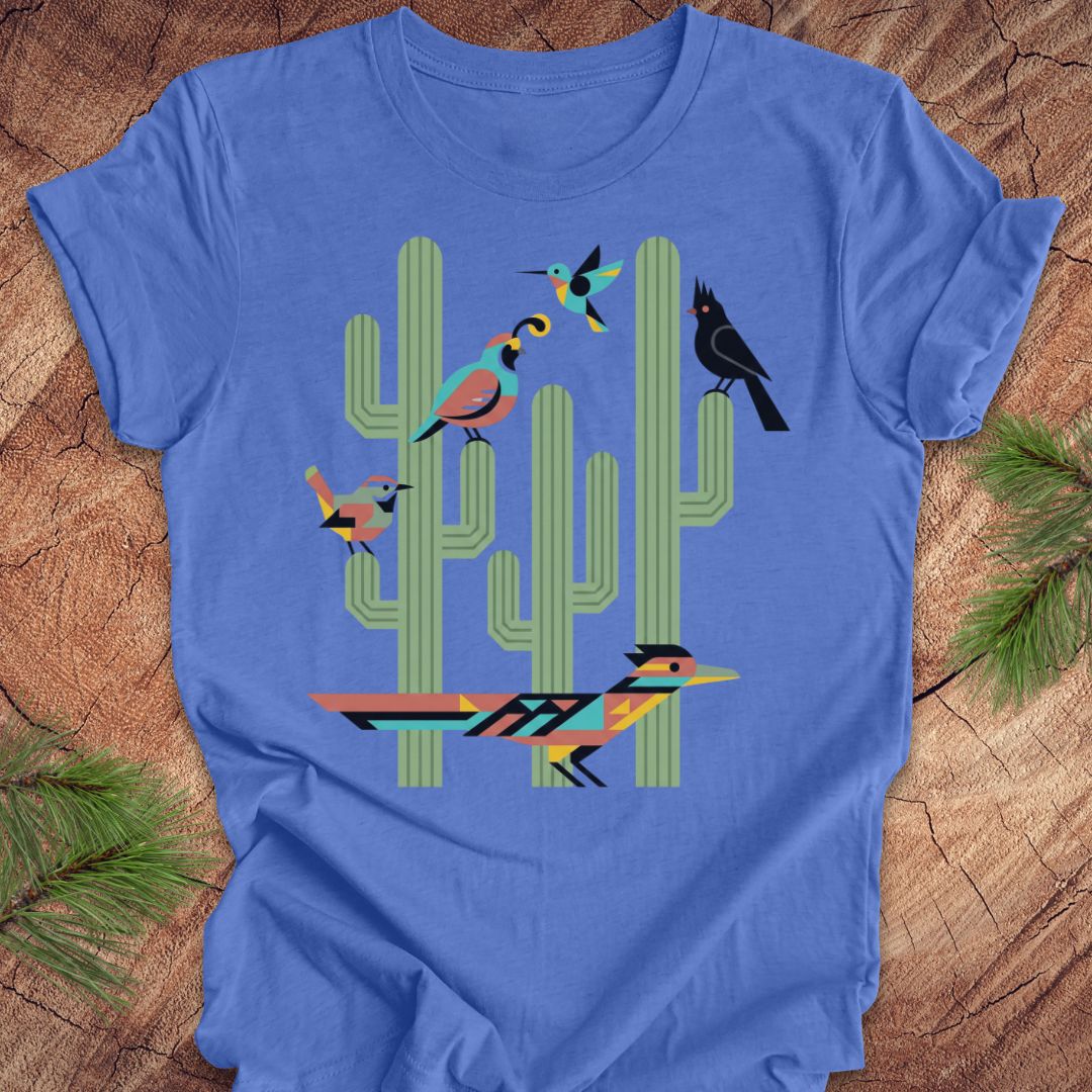 A blue t-shirt with a colorful graphic print of stylized birds perched on green cacti, laid flat on a rustic wood background with pine needles.