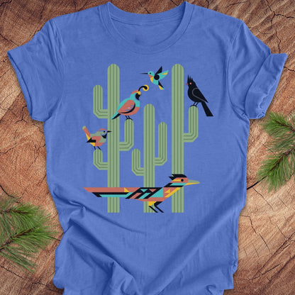 A blue t-shirt with a colorful graphic print of stylized birds perched on green cacti, laid flat on a rustic wood background with pine needles.