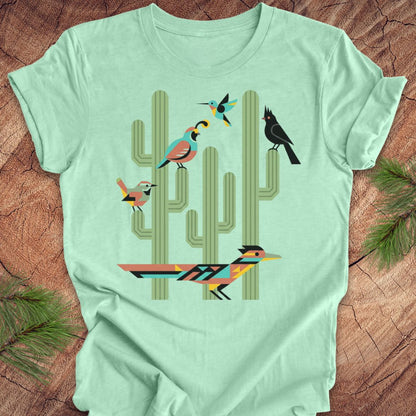 A mint green short-sleeve t-shirt with a colorful graphic print of stylized birds perched on a saguaro cactus, laid flat on a rustic wood background with pine needles.