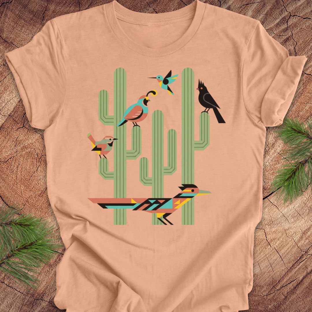 A peach t-shirt with a colorful graphic print of stylized birds perched on green cacti, laid flat on a rustic wood background with pine needles.