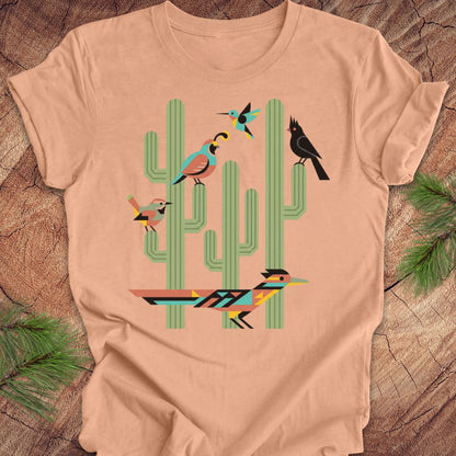 A peach t-shirt with a colorful graphic print of stylized birds perched on green cacti, laid flat on a rustic wood background with pine needles.