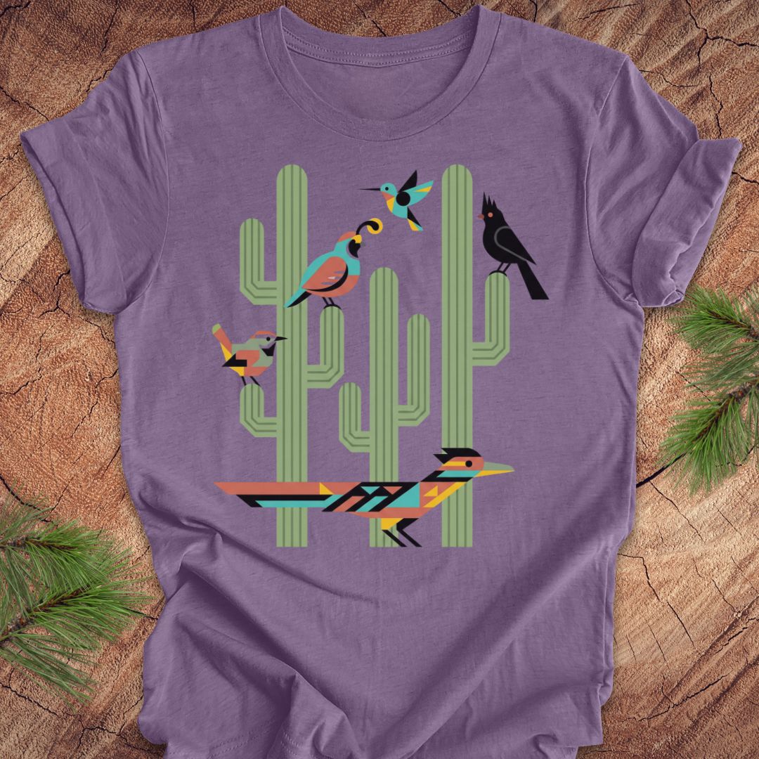 A purple t-shirt with a colorful graphic print of stylized birds perched on green cacti, laid flat on a rustic wood background with pine needles.