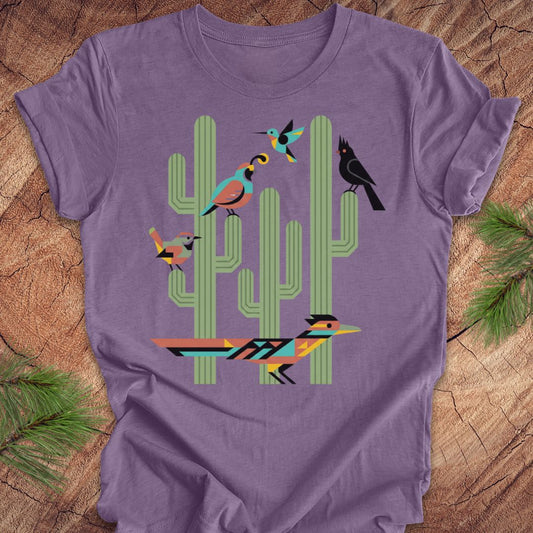 A purple t-shirt with a colorful graphic print of stylized birds perched on green cacti, laid flat on a rustic wood background with pine needles.