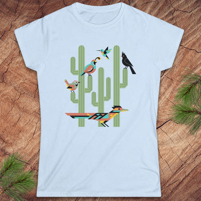 A light blue short-sleeve crewneck t-shirt featuring a colorful graphic of stylized birds perched on green cacti, laid flat on a rustic wood background with pine needles in the corners.