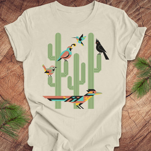 A beige t-shirt with a colorful graphic print of stylized birds perched on green cacti, laid flat on a rustic wood background with pine needles.