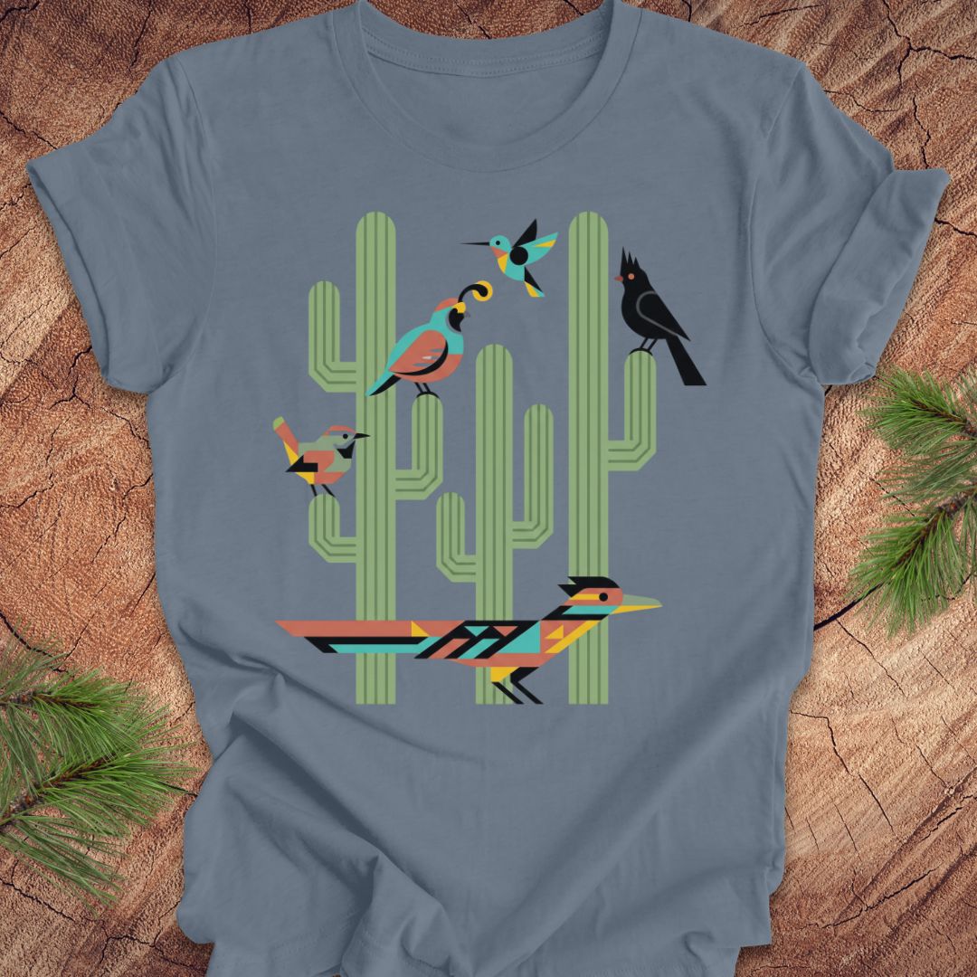 A slate blue t-shirt with a colorful graphic print of stylized birds perched on green cacti, laid flat on a rustic wood background with pine needles.