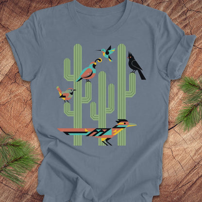 A slate blue t-shirt with a colorful graphic print of stylized birds perched on green cacti, laid flat on a rustic wood background with pine needles.