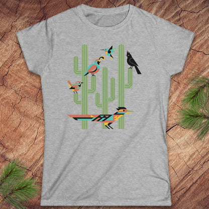 A heather gray short-sleeve t-shirt with a colorful graphic print of stylized birds perched on green cacti, laid flat on a textured wood background with pine needles in the corners.