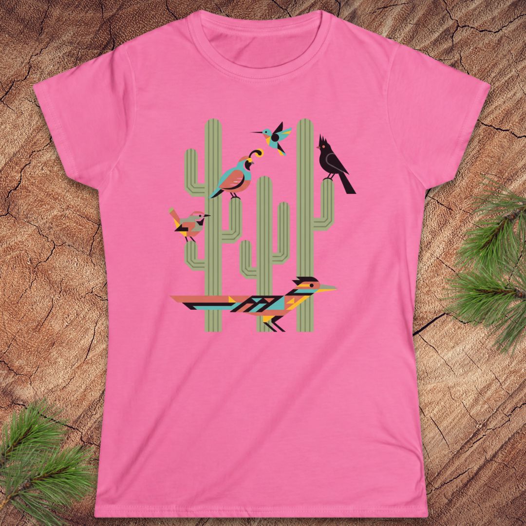 A pink short-sleeve crewneck t-shirt featuring a colorful graphic of stylized birds perched on green cacti, laid flat on a rustic wood background with pine needles in the corners.