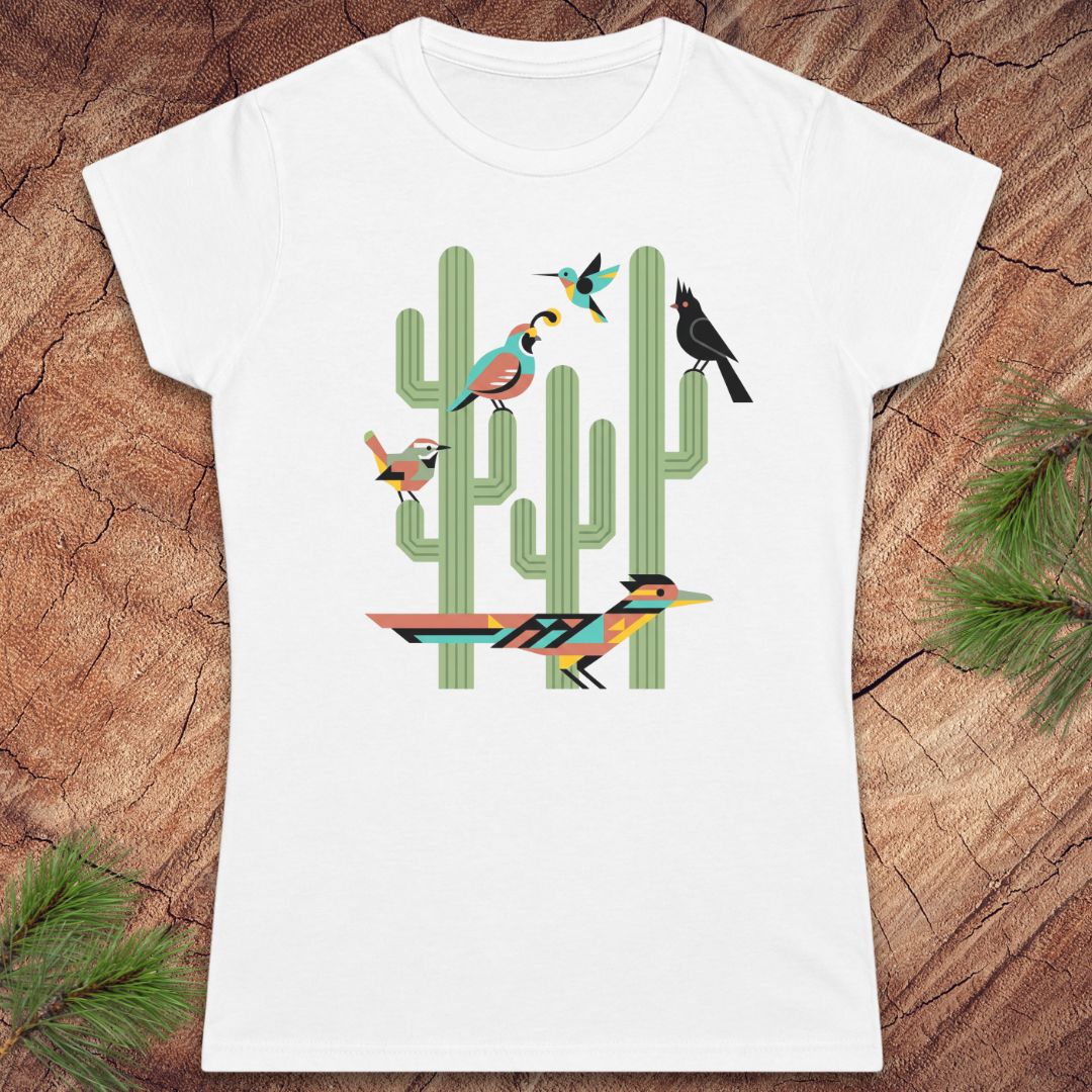 A white short-sleeve crewneck t-shirt featuring a colorful graphic of stylized birds perched on green saguaro cacti, displayed flat on a rustic wood background with pine needles.