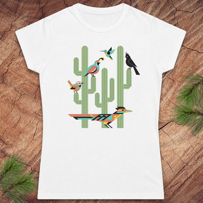 A white short-sleeve crewneck t-shirt featuring a colorful graphic of stylized birds perched on green saguaro cacti, displayed flat on a rustic wood background with pine needles.