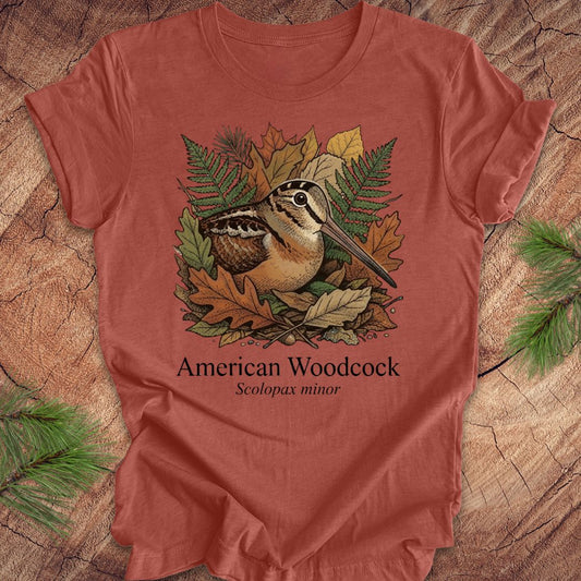Clay colored t-shirt with an illustration of an American Woodcock and foliage on a wooden surface.