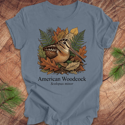 Blue t-shirt with an illustration of an American Woodcock and autumn leaves on a wooden surface.
