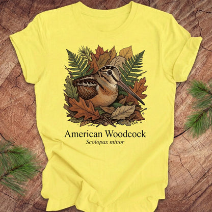 Yellow t-shirt with an illustration of an American Woodcock and leaves on a wooden surface.