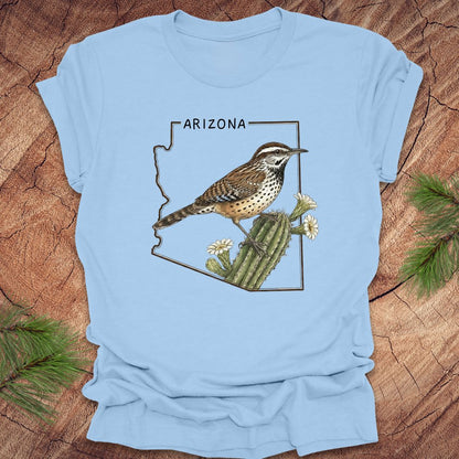 Baby blue t-shirt with Arizona state outline with cactus wren and cactus flower design on a wooden surface
