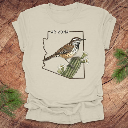 Beige t-shirt with Arizona state outline with cactus wren and cactus flower design on a wooden surface