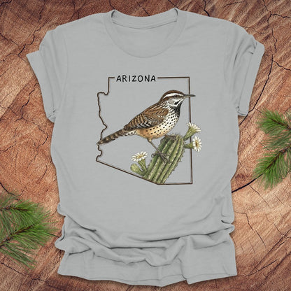 Silver t-shirt with Arizona state outline with cactus wren and cactus flower design on a wooden surface
