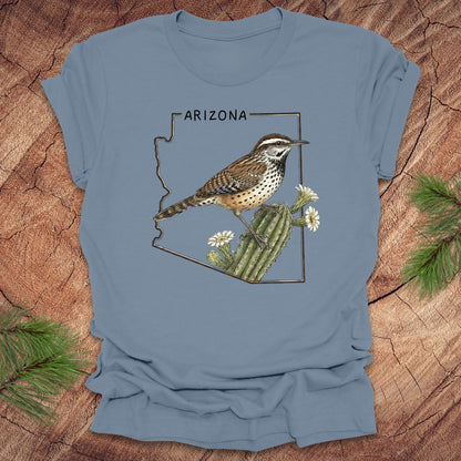 Steel blue t-shirt with Arizona state outline with cactus wren and cactus flower design on a wooden surface