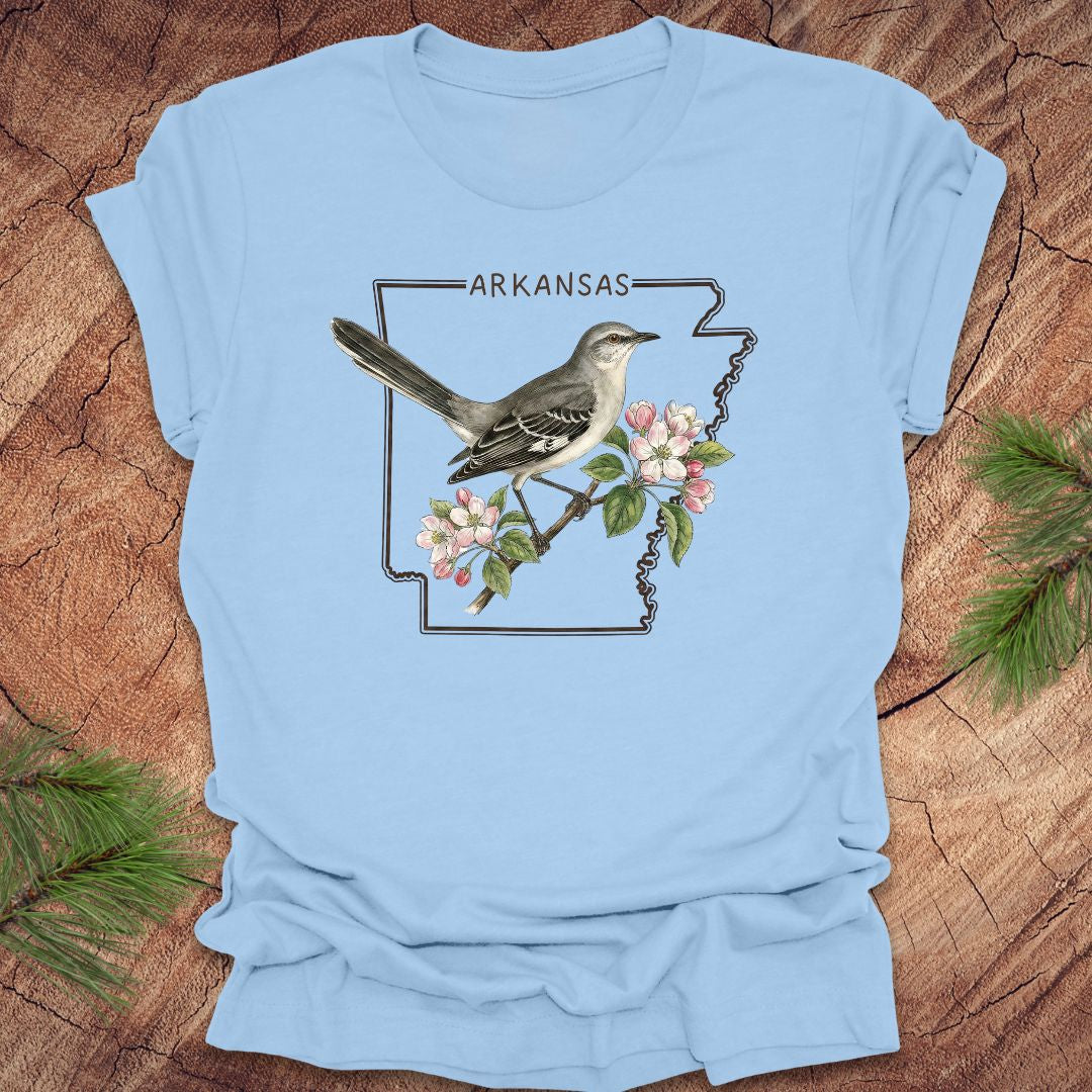 Light blue t-shirt with a bird and floral design on Arkansas outline on a wooden surface.