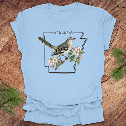 Light blue t-shirt with a bird and floral design on Arkansas outline on a wooden surface.