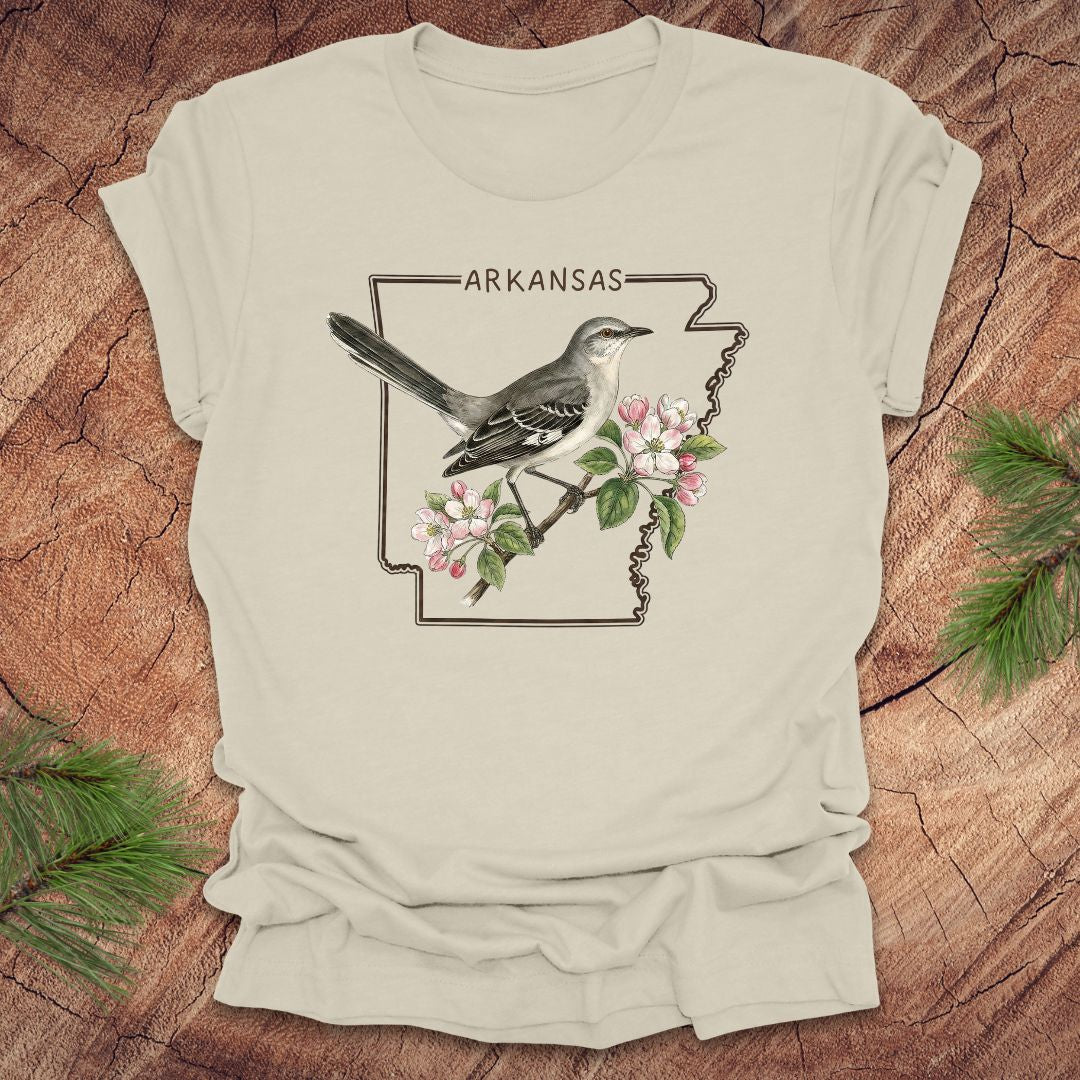 Beige t-shirt with Arkansas state design featuring a bird and flowers on a wooden surface.